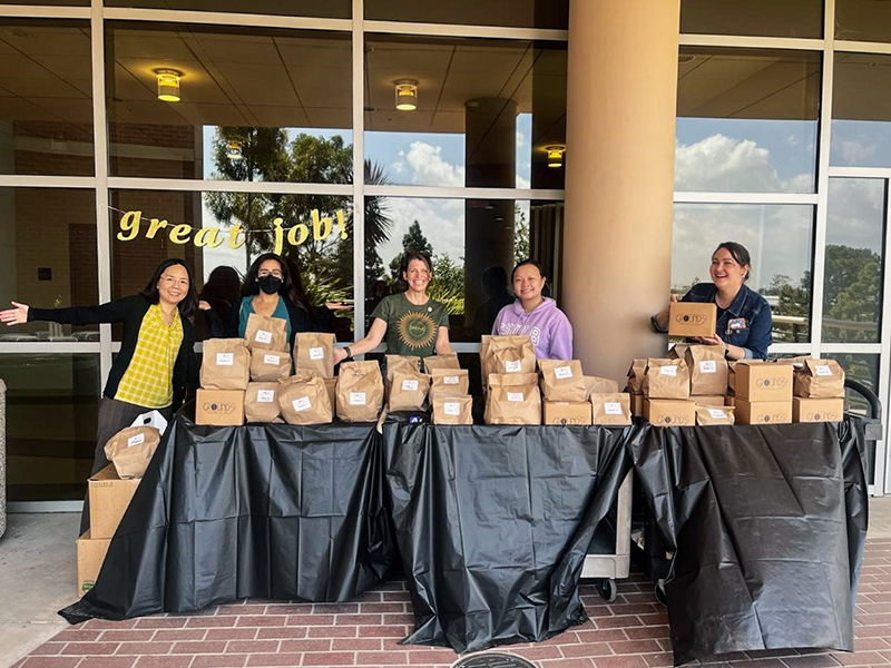 Outdoor photo of 5 people. (Left  to Right): The set up team! Panadda Marayong, Sarah Velasco, Kelly Young, Cindy Su, and Nicole Streicker.  A banner that says “Great Job” hangs on the window behind them.